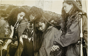 Four young musicians form a tight circle, heads bowed, during a performance with a microphone stand nearby.