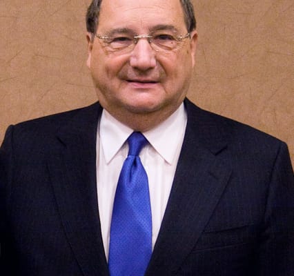 Smiling man in a dark suit and blue tie posing against a beige wall