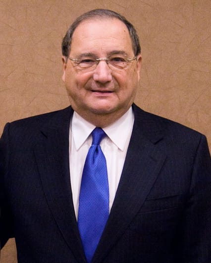 Smiling man in a dark suit and blue tie posing against a beige wall