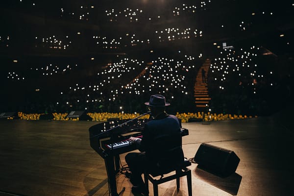 Pianist wearing a hat playing a grand piano on a dimly lit stage, with small bright audience lights in the background.