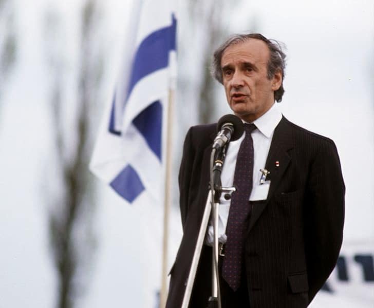 Man in a dark suit speaks into a microphone at an outdoor event, with a blue-and-white flag in the background.