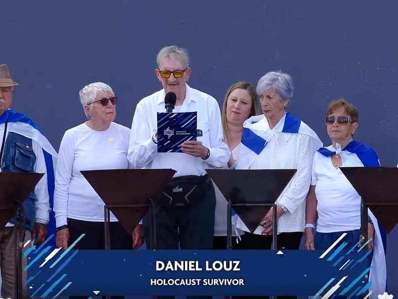 Man speaks into a microphone at a podium, flanked by six adults on stage, all wearing white tops and blue scarves; banner shows 'DANIEL LOUZ' and 'HOLOCAUST SURVIVOR'.