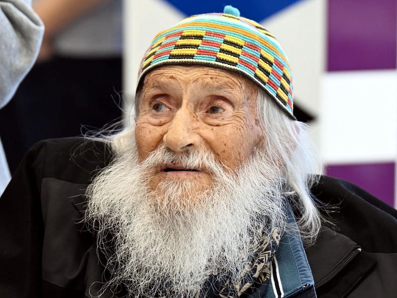 Older man with a long white beard wearing a colorful knit cap and dark jacket, looking off to the left.